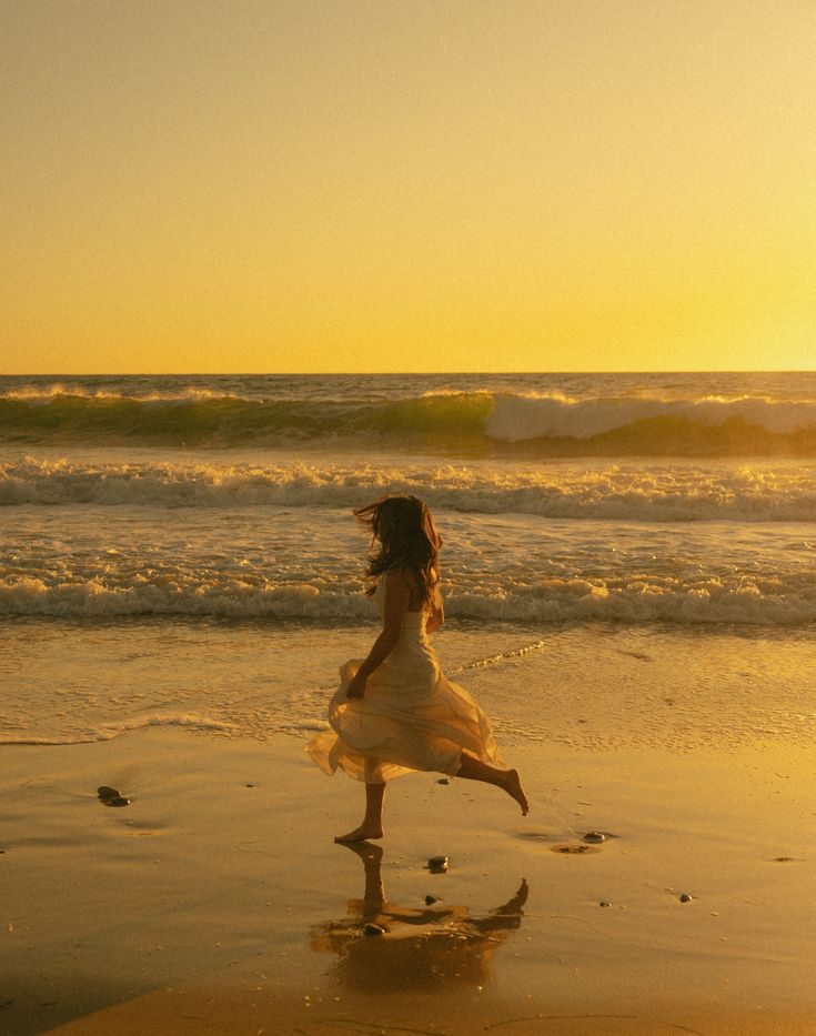 A girl in a flowing light dress running barefoot along the wet sand at the water's edge, bathed in golden light as waves roll in at sunset